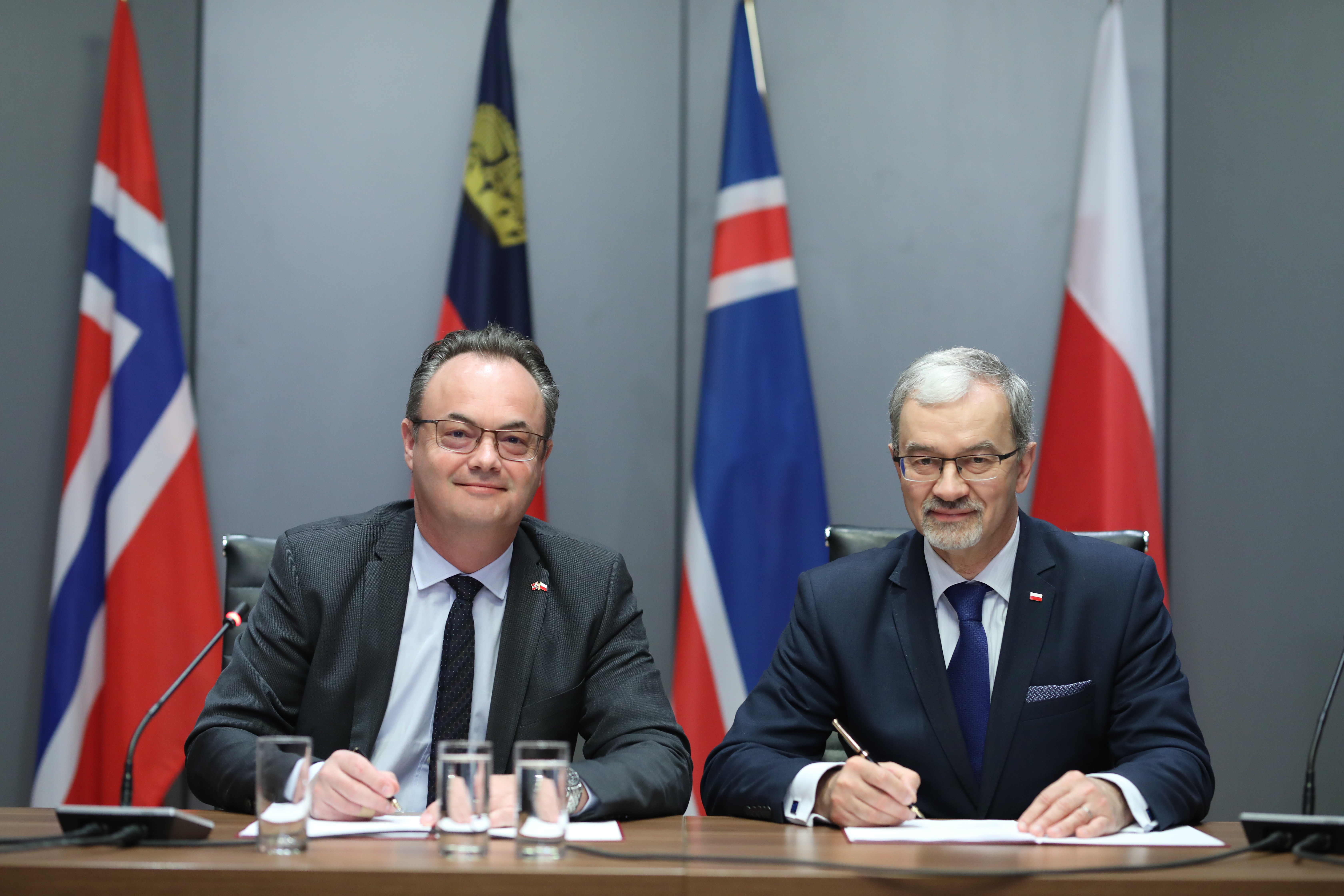 Minister Inwestycji i Rozwoju Jerzy Kwieciński i Ambasador Norwegii Olav Myklebust siedzą przy stole, w dłoniach trzymają pióra. W tle od prawej stoją flagi Polski, Islandii, Liechtensteinu i Norwegii.